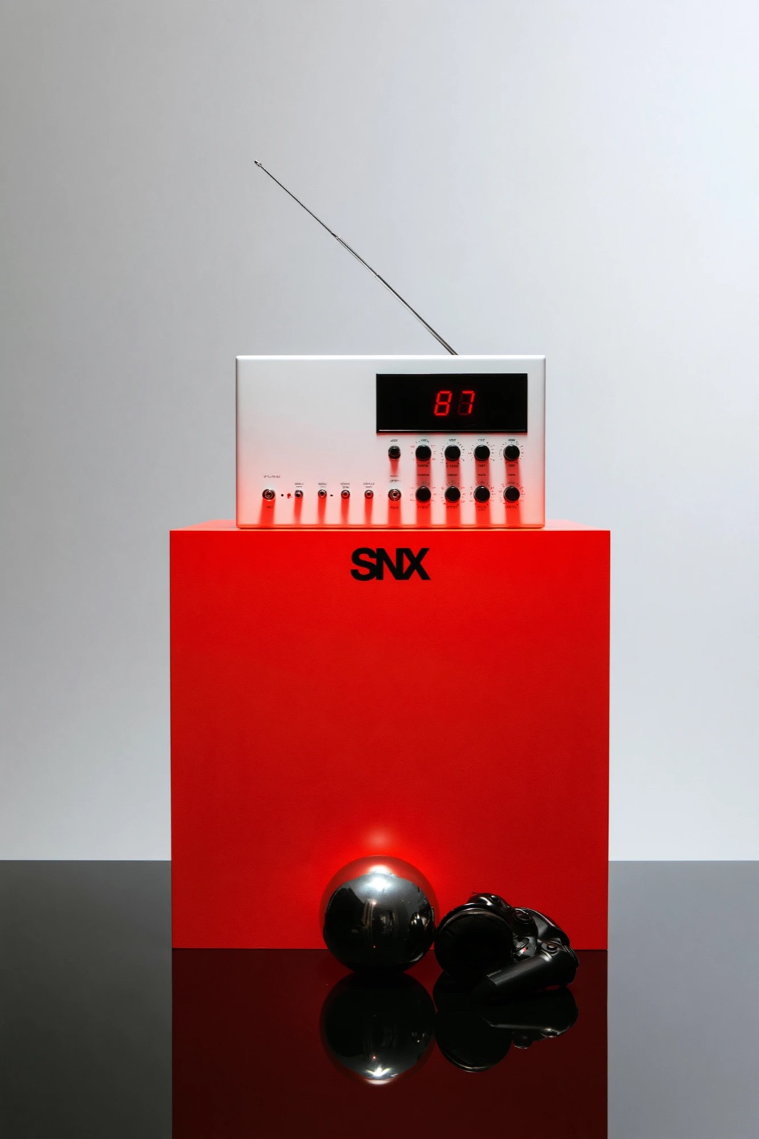 A white radio with a red display sits on a red SNX block, beside a chrome ball and headphones