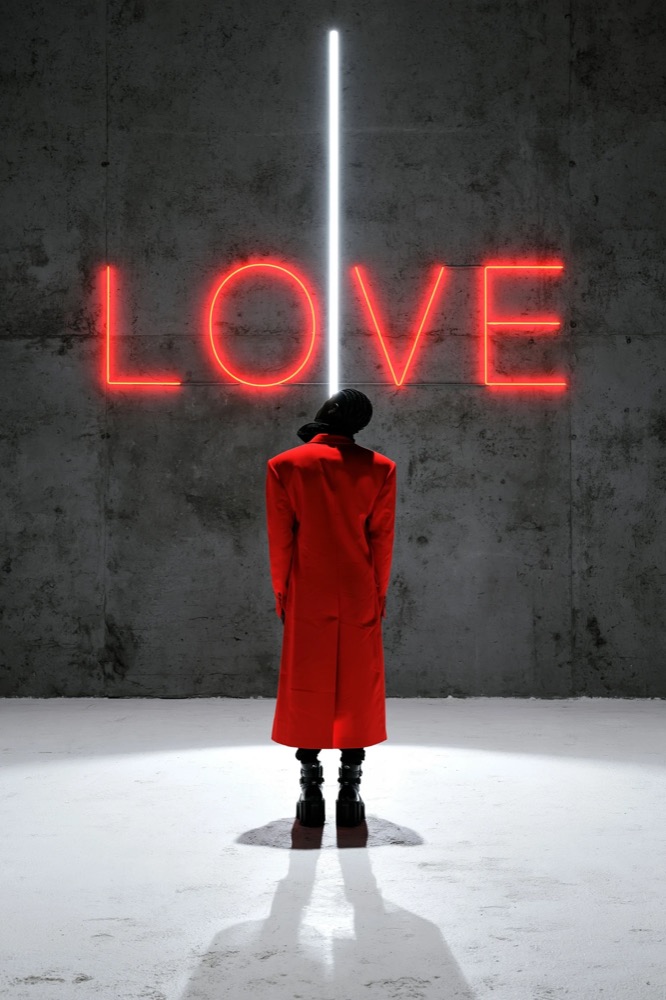 A figure in a long red coat stands with their back to us under a red neon LOVE sign on a concrete wall