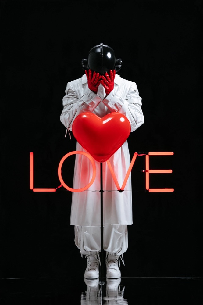 A figure in white with a black helmet covers their face with red gloves behind a red heart and a neon LOVE sign