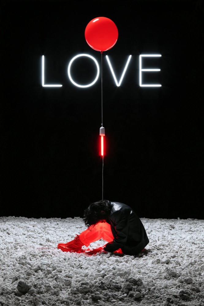 A figure curled on white gravel beneath a red balloon and a white neon LOVE sign
