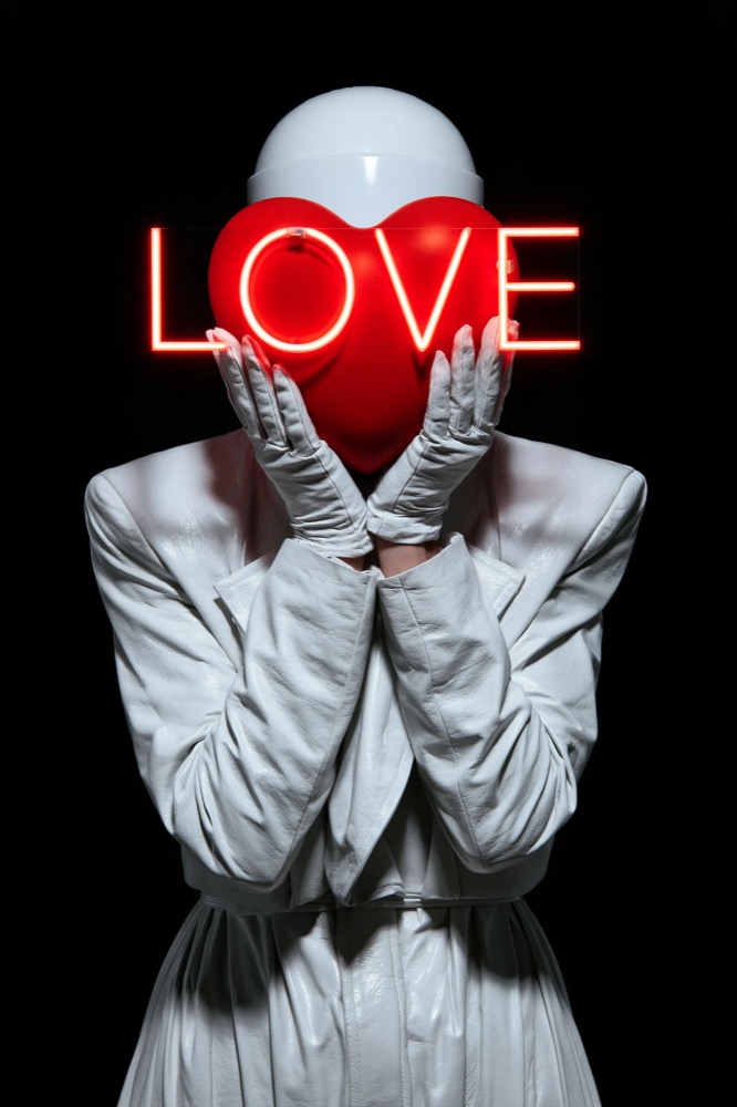 A figure in white leather holds a red heart and a neon LOVE sign in front of a white helmet