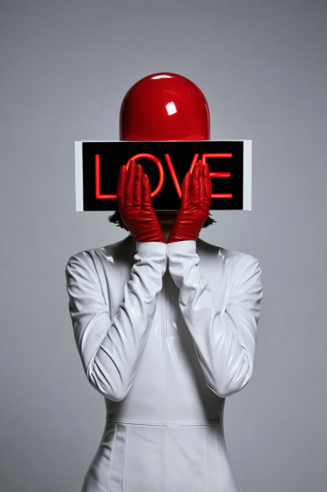 A figure in white with a red dome helmet hides behind a LOVE sign held in red gloves