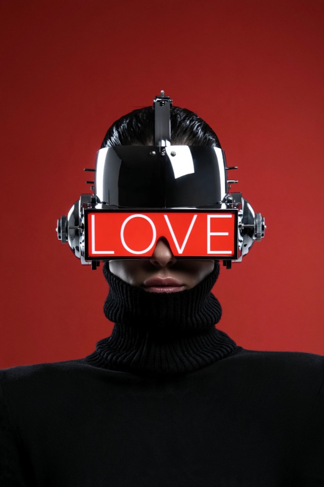 A figure in a black turtleneck wears a chrome helmet with a red LOVE visor against a red wall