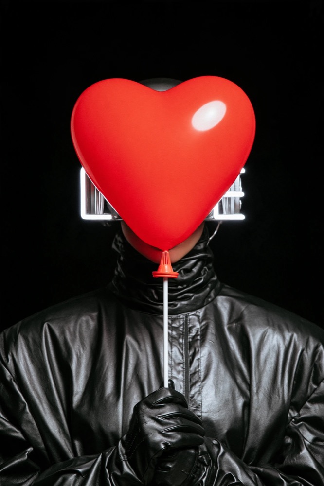 A figure in black leather holds a red heart balloon in front of their face