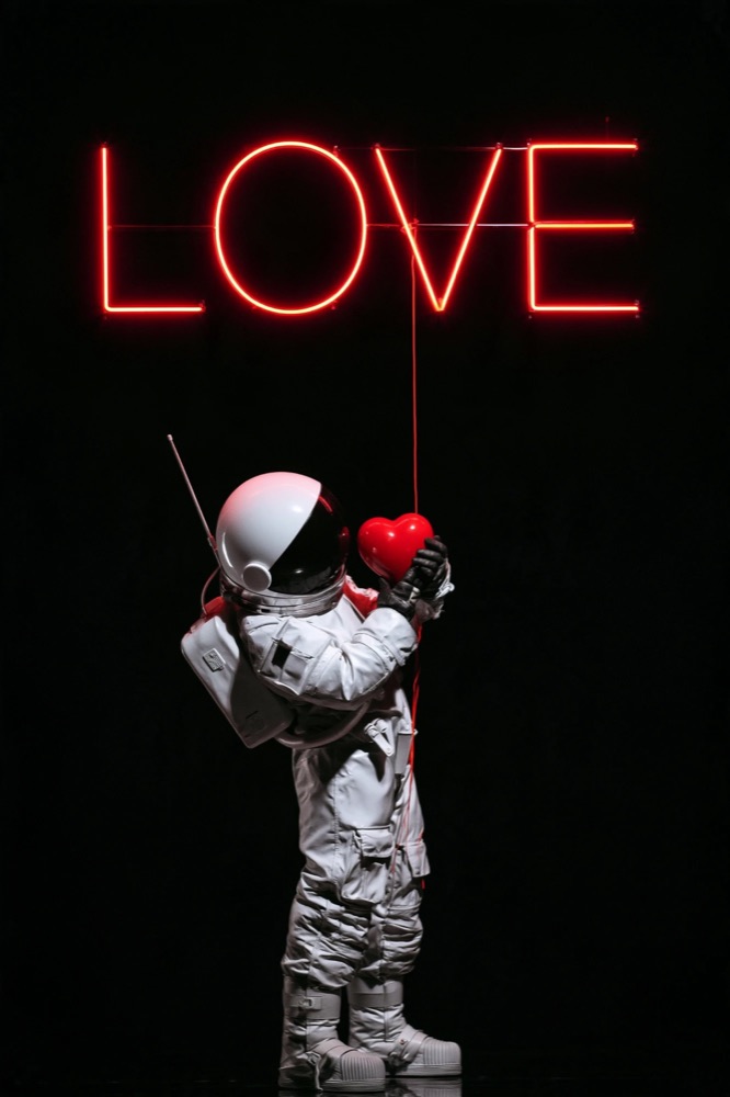 A small astronaut in white hugs a red heart under a red neon LOVE sign
