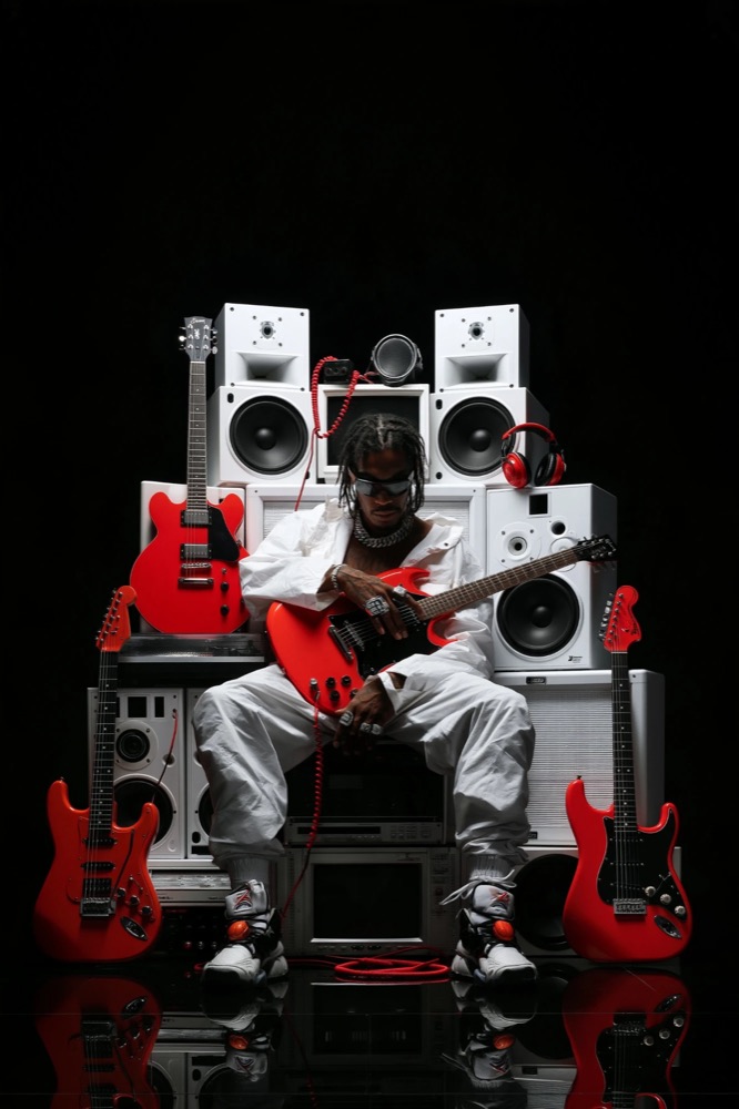 A man in white sits on a stack of white speakers holding a red guitar, with three more red guitars around him