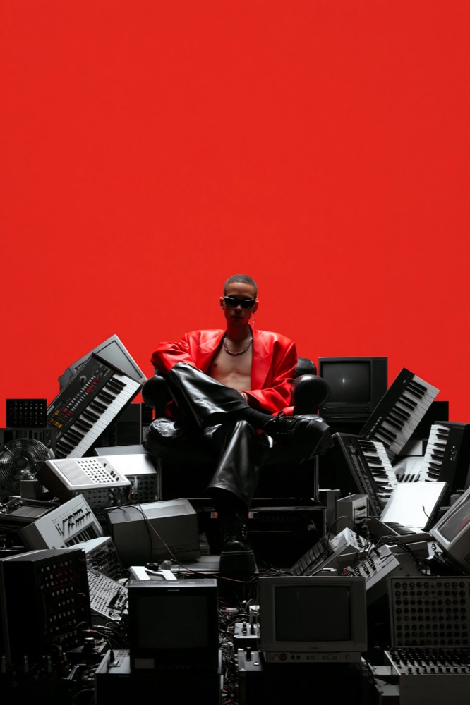 A man in a red leather jacket sits in an armchair on a heap of keyboards and old monitors, against a red wall