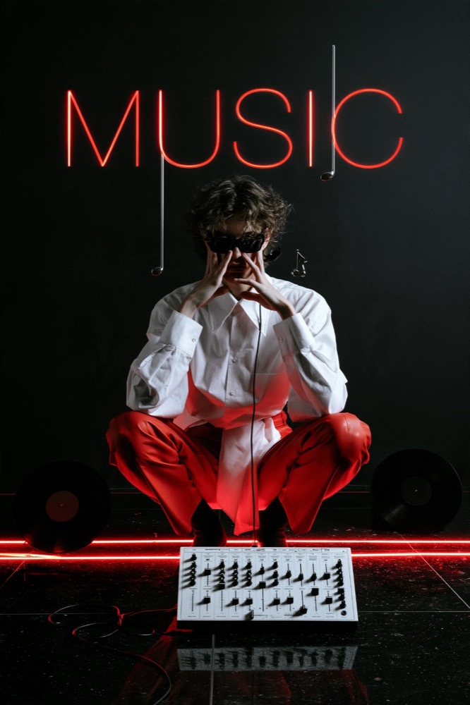 A figure in a white shirt and red trousers crouches under a red neon sign reading MUSIC, a mixer on the floor