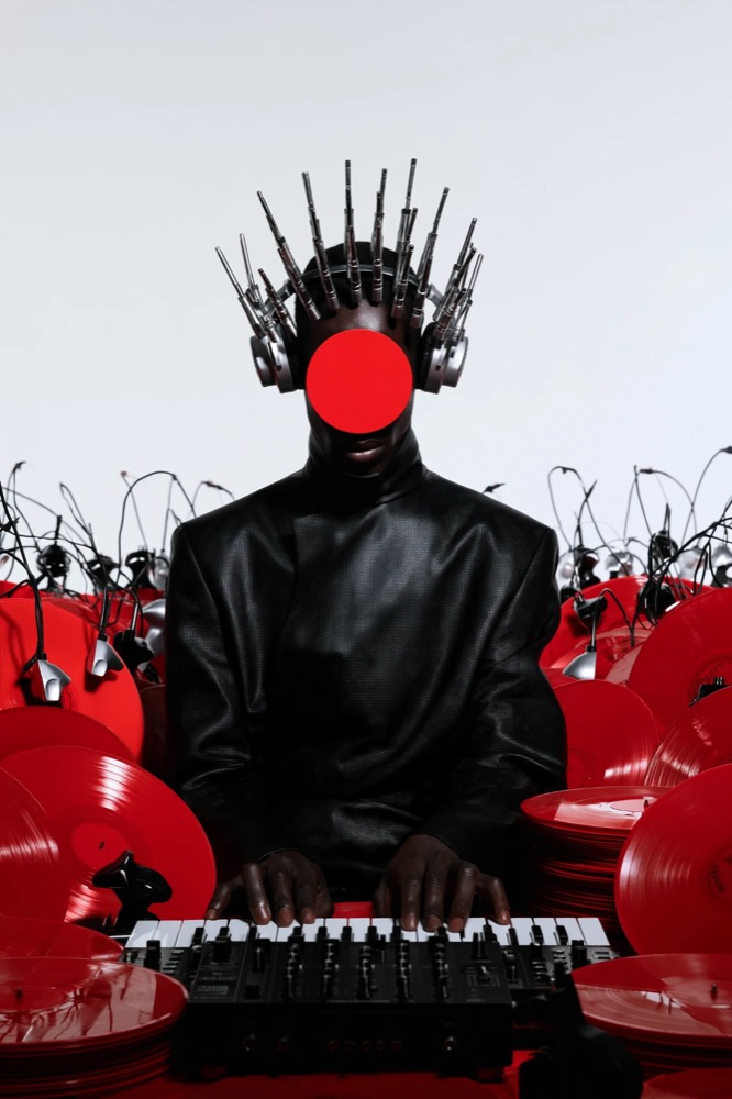 A figure in black with a red disc for a face and a crown of audio plugs plays a keyboard among red vinyl records