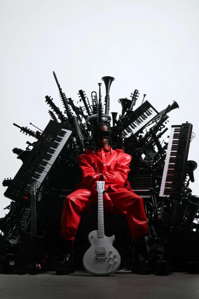 A figure in red sits on a throne made of black keyboards, horns and guitars, holding a white guitar