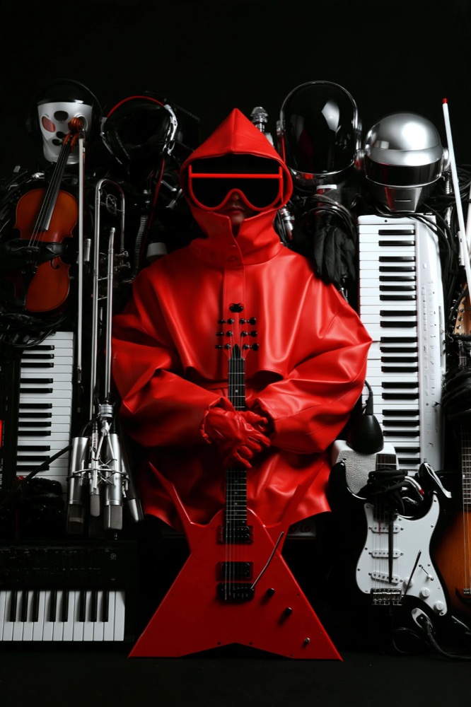 A hooded figure in red with red goggles holds a red V-shaped guitar among keyboards, helmets and instruments