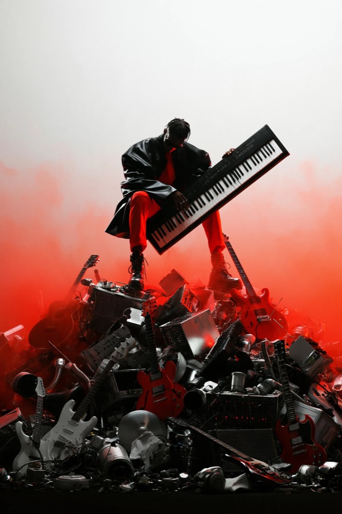 A man in a black coat and red suit stands on a mountain of guitars and amps holding a keyboard in red smoke