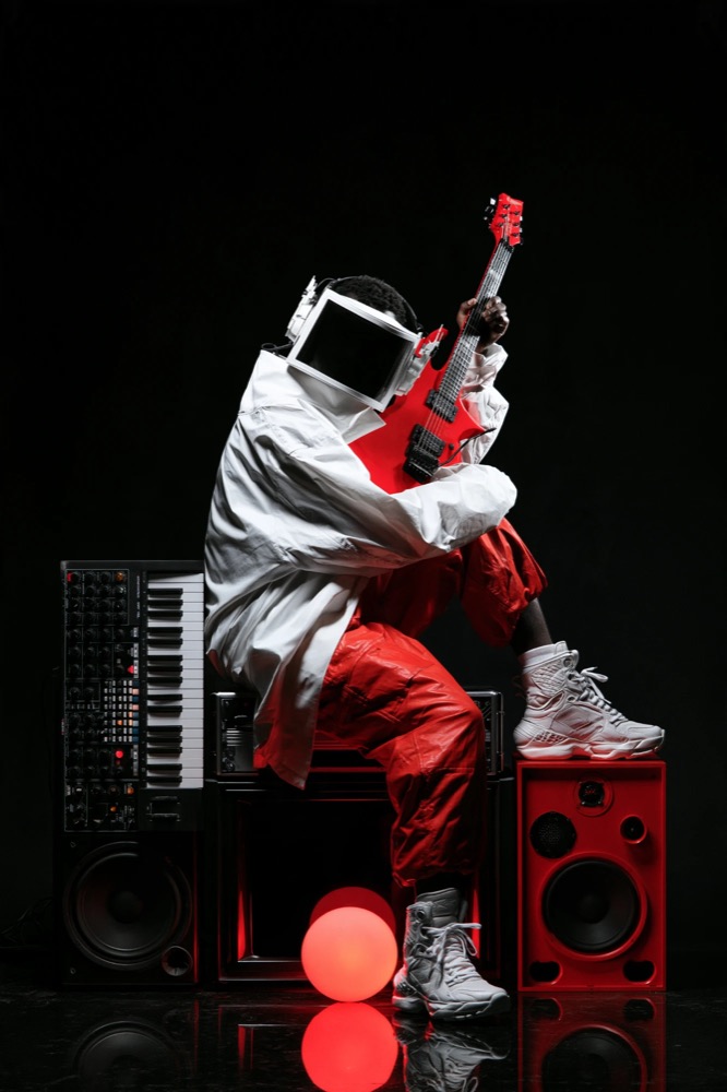 A figure in white with a monitor for a head hugs a red guitar, sitting on speakers beside a glowing red ball