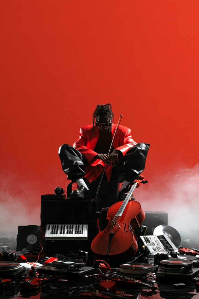 A man in a red jacket crouches on speakers behind a cello, surrounded by turntables and records in red fog